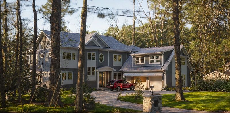 Front view of a large gray two story house with attached garage, driveway, red SUV, and tall trees around.