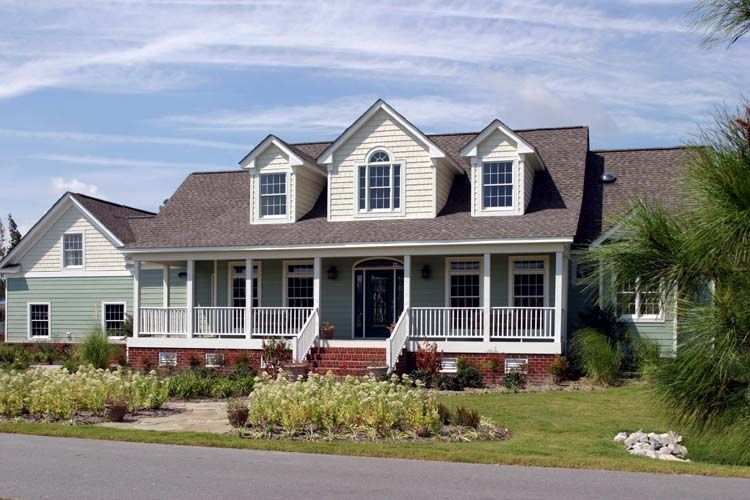 Front view of a white single-story house with dormer windows, front porch, landscaped garden, and paved road up.