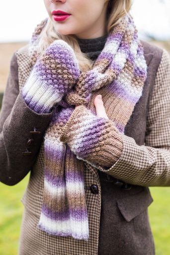 A woman is wearing a knitted scarf and matching wristwarmers in a variegated purple, cream and brown yarn.