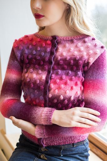 A woman is wearing a variegated pink and purple knitted cardigan with bobbles on the front.