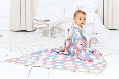 A baby is wrapped in a crocheted blanket made up oh pink, blue, purple and cream hexagons and joined together with green.
