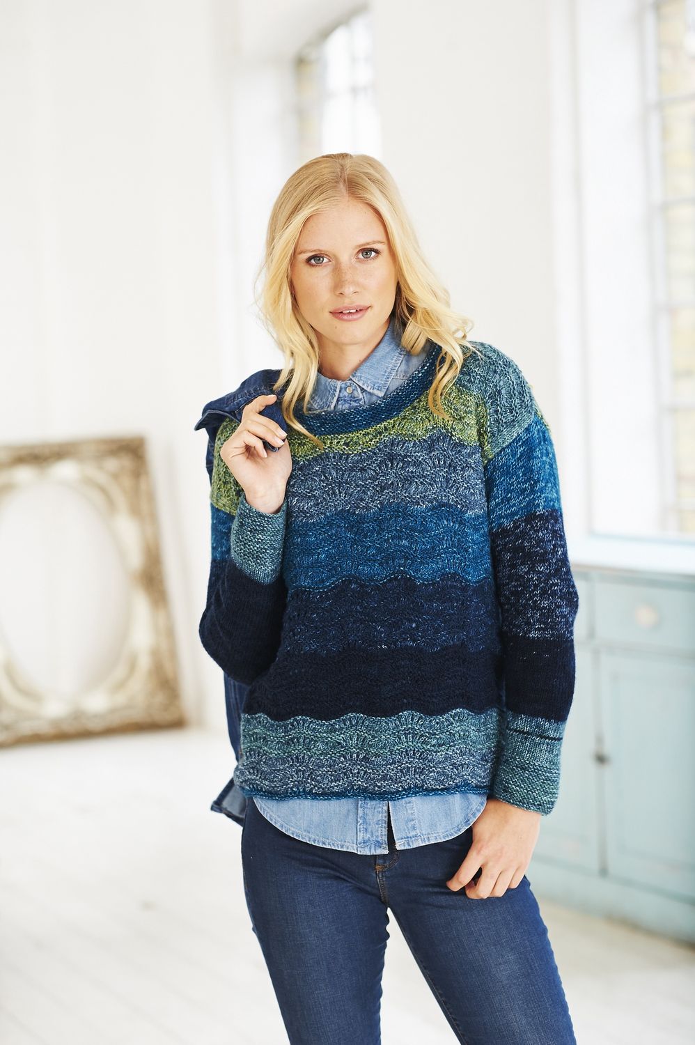 A woman is wearing a knitted sweater in a variegated blue and green yarn.