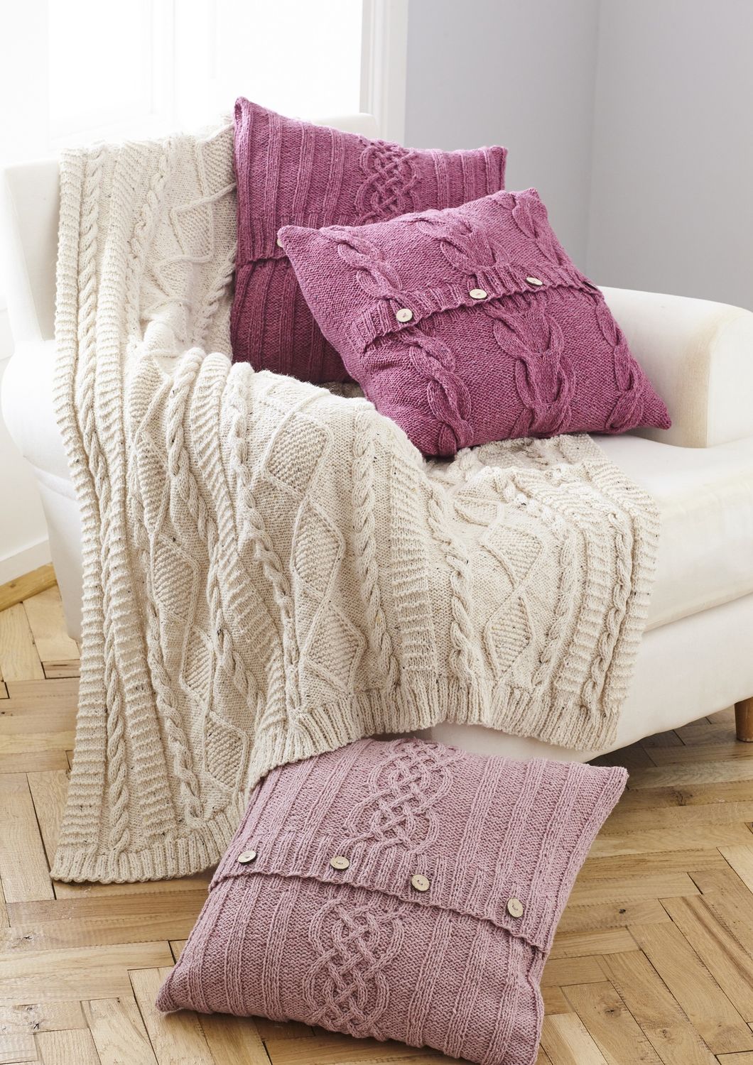 A cream cable knit blanket with 3 pink cable knit cushions.