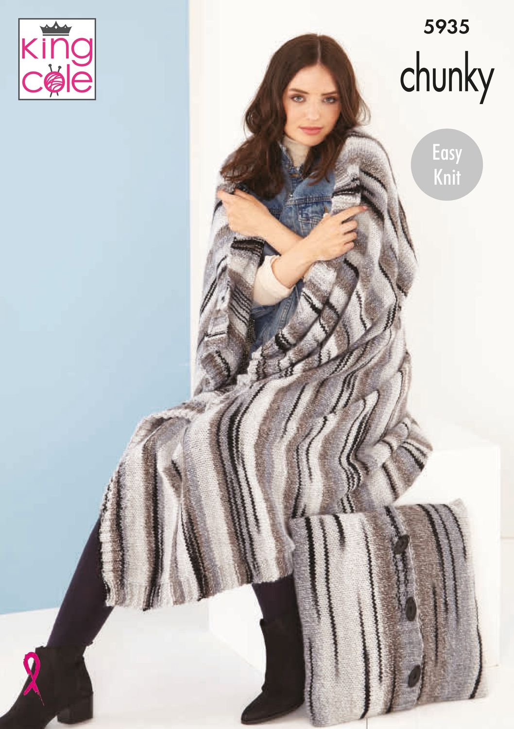 A woman is sat down with a variegated knitted blanket in shades of cream and brown with random thin black stripes throughout draped around her shoulders. A matching cushion is sat on the floor next to her.