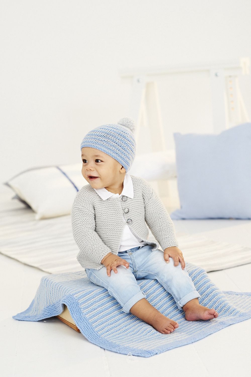 A baby is sitting on a blue knitted blanket while wearing a grey knitted cardigan and a blue knitted hat.