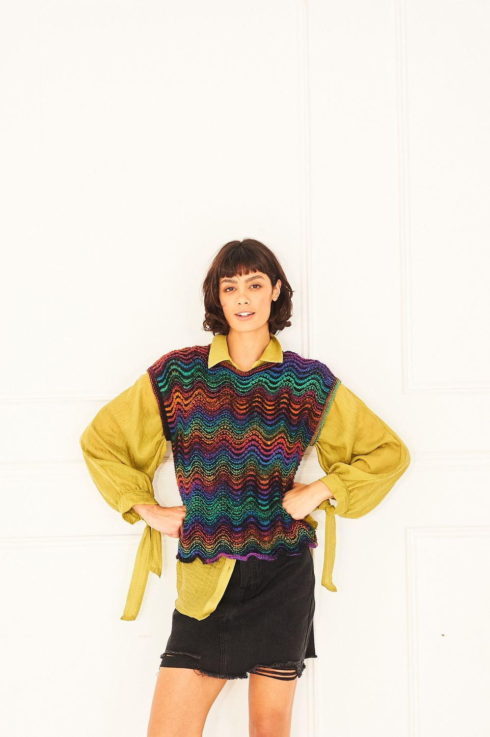A woman is wearing a knitted tank top in shades of black, red, orange, green and blue and purple.
