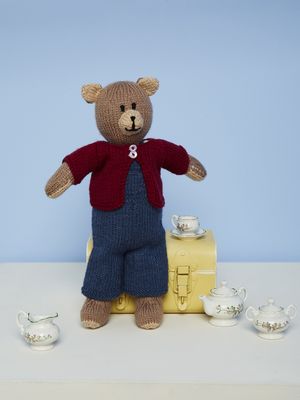 A brown knitted bear is wearing blue dungarees and a red cardigan.