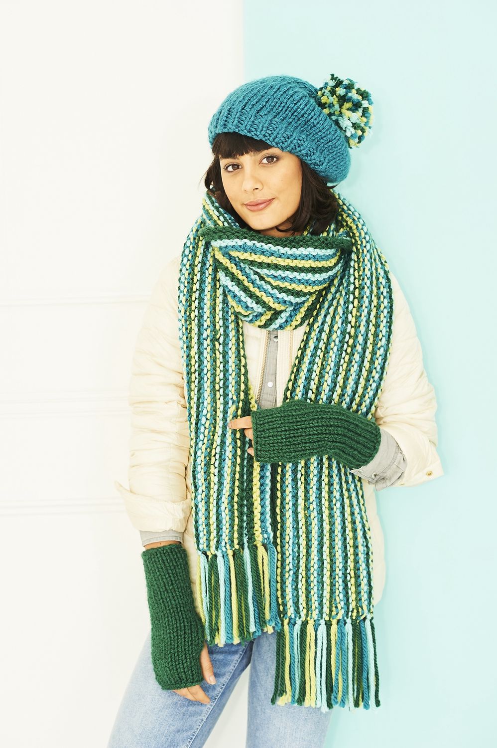 A woman is wearing a green and blue stripy knitted scarf and a blue knitted hat with a blue and green pompom.