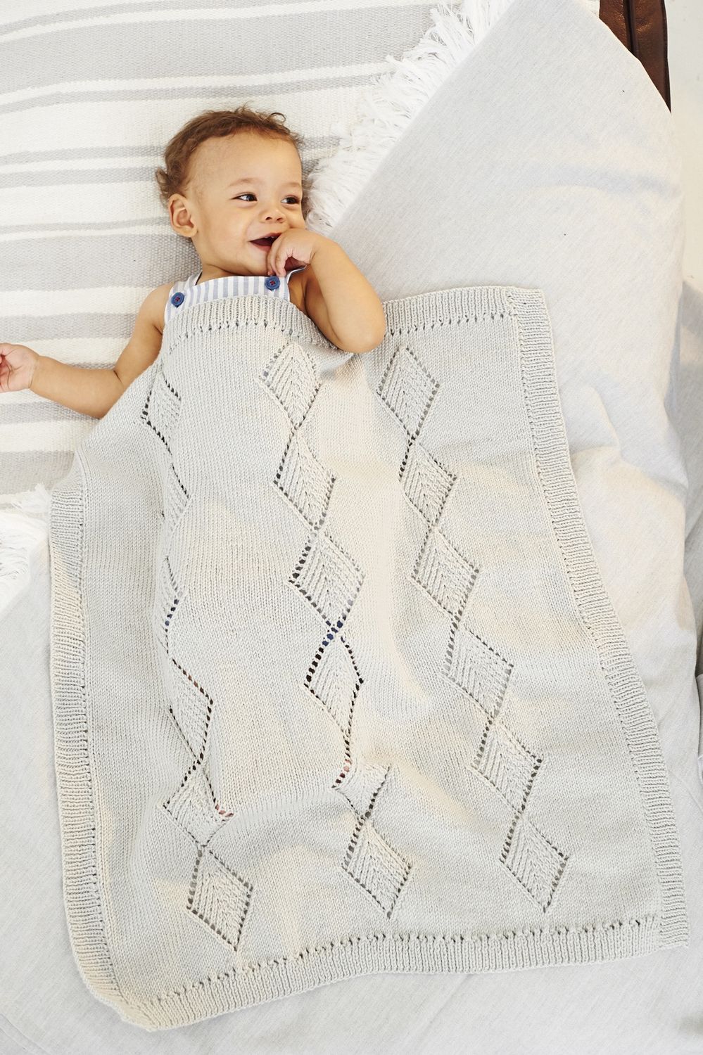 A baby is under a grey knitted blanket with a lace diamond pattern.