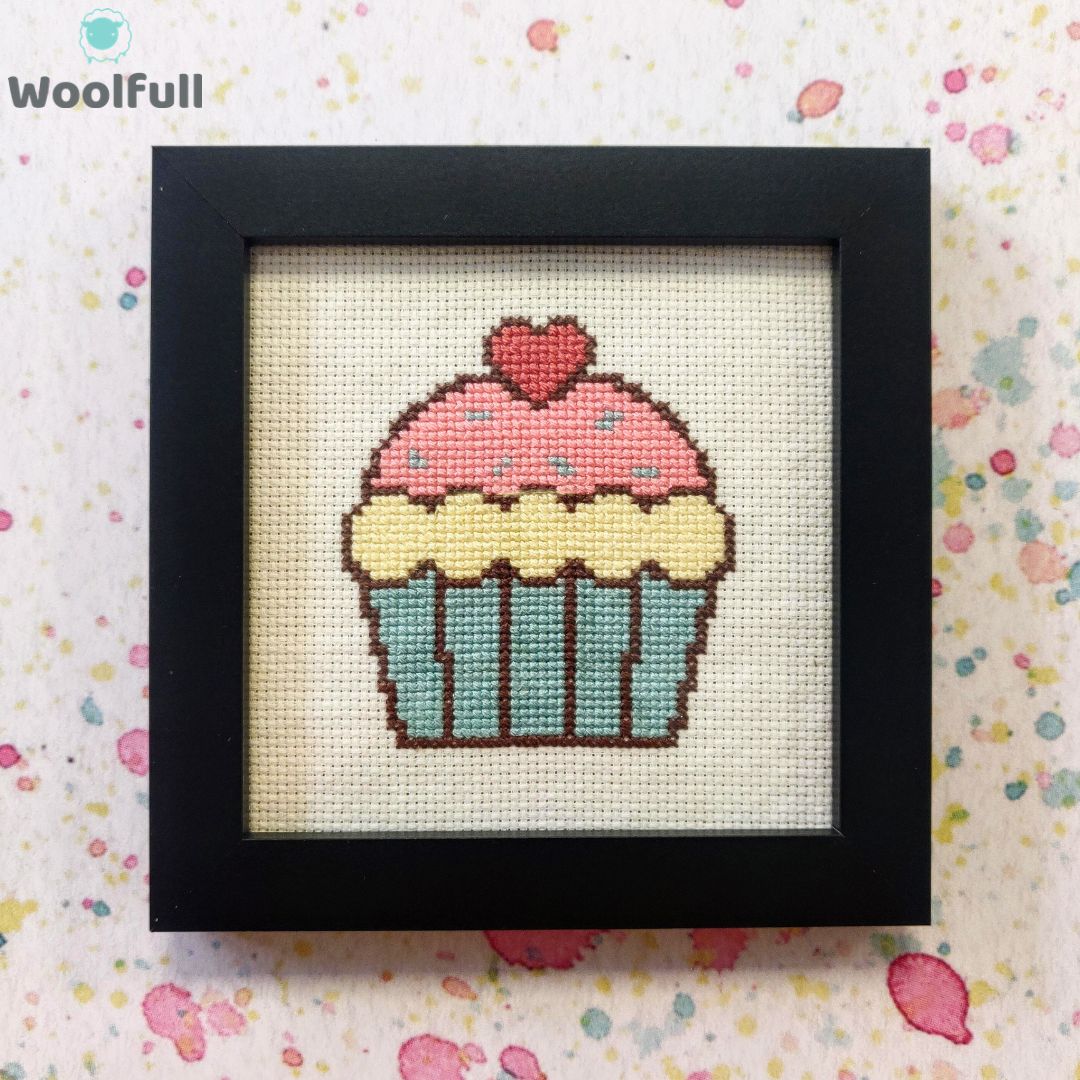 A framed cross stitch with a design of a cartoon style cupcake.