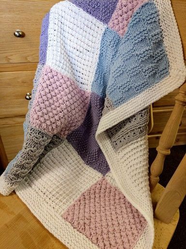 A knitted blanket made up of different textured squares in white, grey, blue, purple and pink.