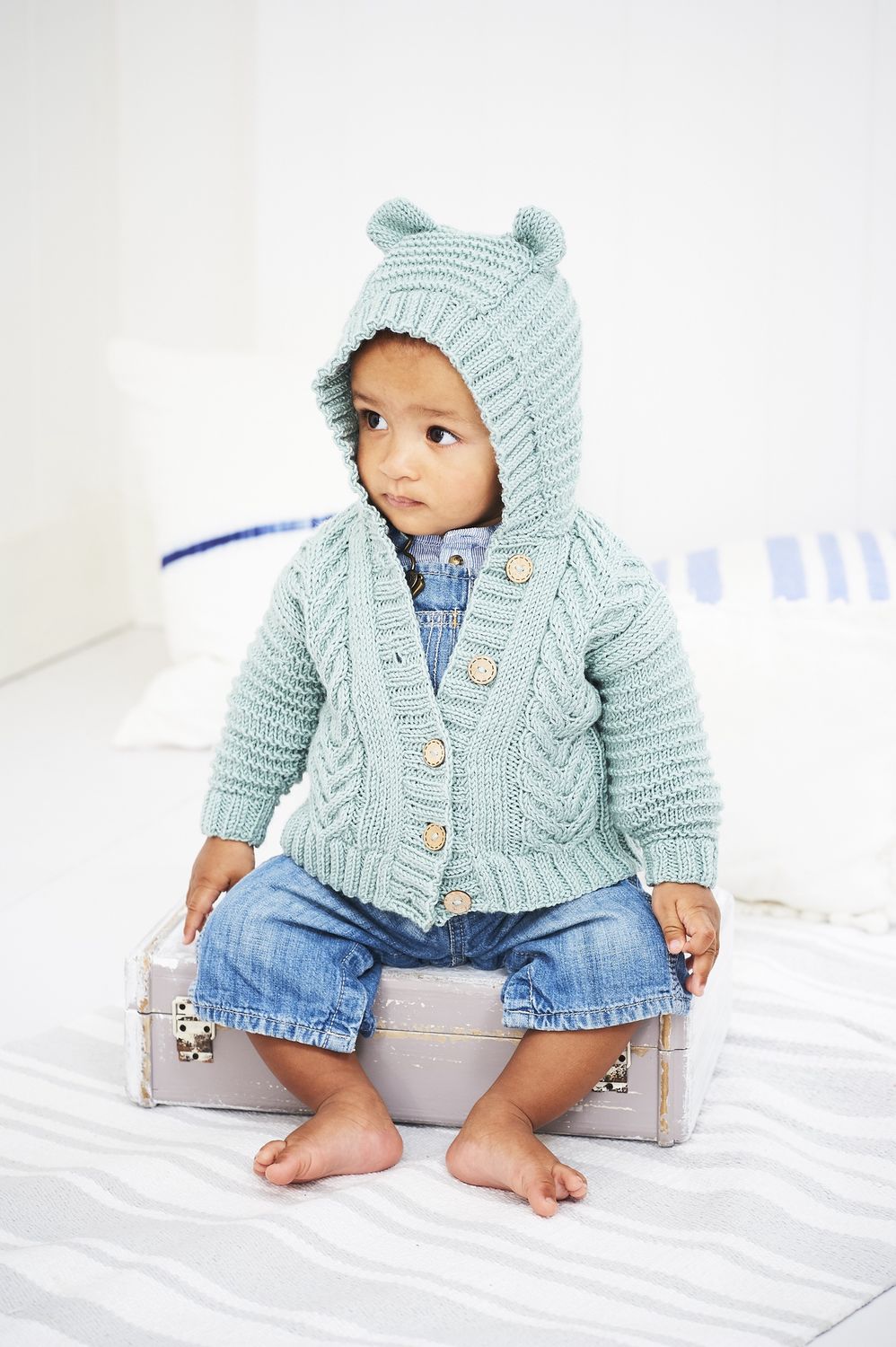 A baby is wearing a pale blue cable knit hooded cardigan.