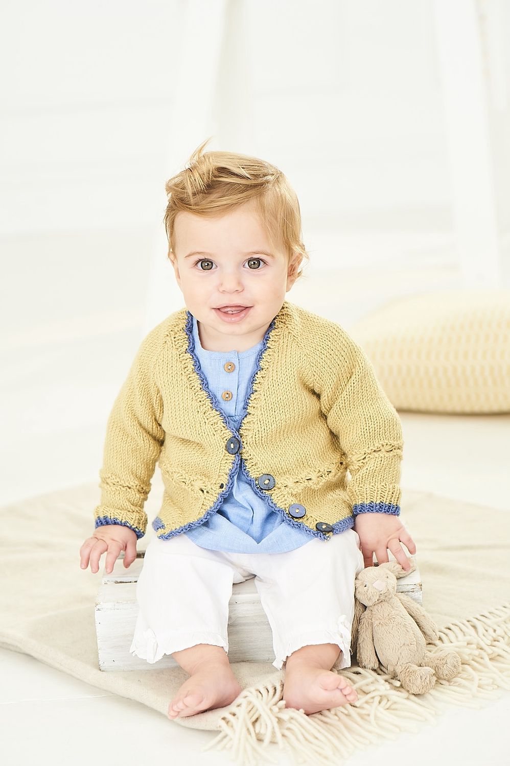 A baby is wearing a yellow knitted cardigan with dark blue edging and buttons.