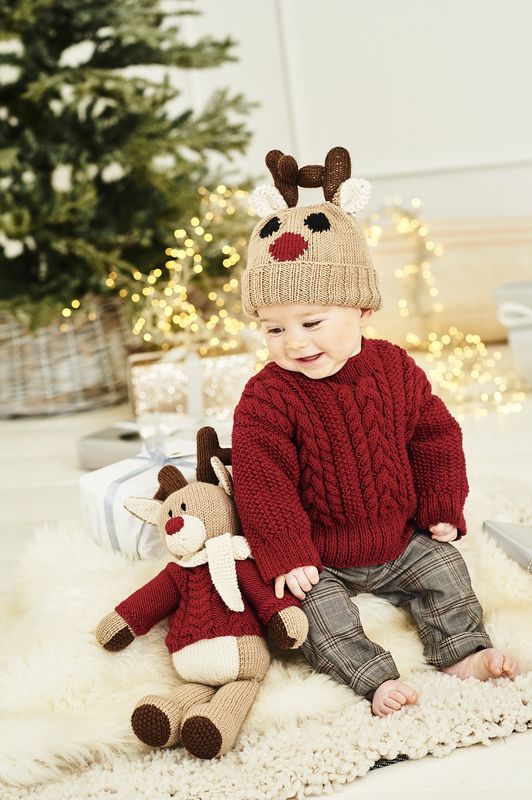 A baby is wearing a red cable knit sweater and a brown reindeer hat, while sitting next to a knitted reindeer toy.