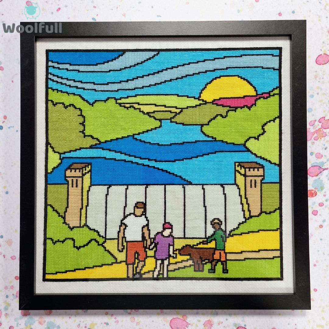 A framed cross stitch with a cartoon style design of Derwent Dam in the Peak District with people and a dog in front.