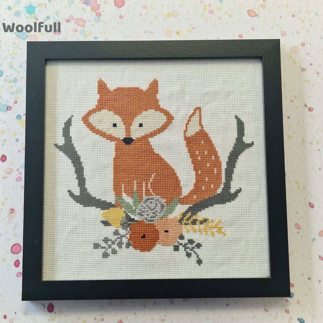 A framed cross stitch with a design of a cartoon style fox alongside branches, leaves and flowers.