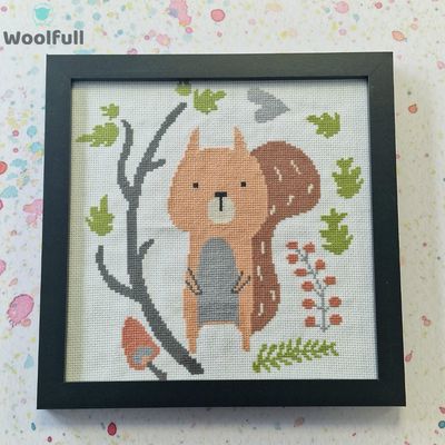 A framed cross stitch with a design of a cartoon style squirrel alongside branches, leaves and flowers.