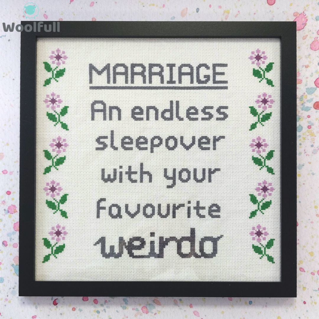 A framed cross stitch with a design that says &#39;marriage, an endless sleepover with your favourite weirdo&#39; in grey and purple and green flowers down each side.