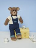 A brown crocheted bear wearing a white top and dark blue dungarees.