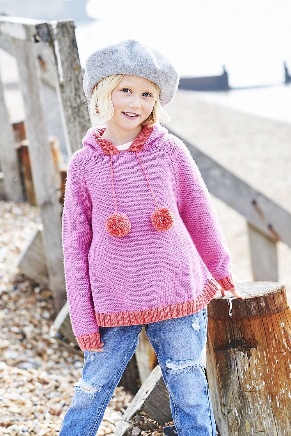 A girl is wearing a pink knitted hoodie, with red pompoms, hood and cuffs.