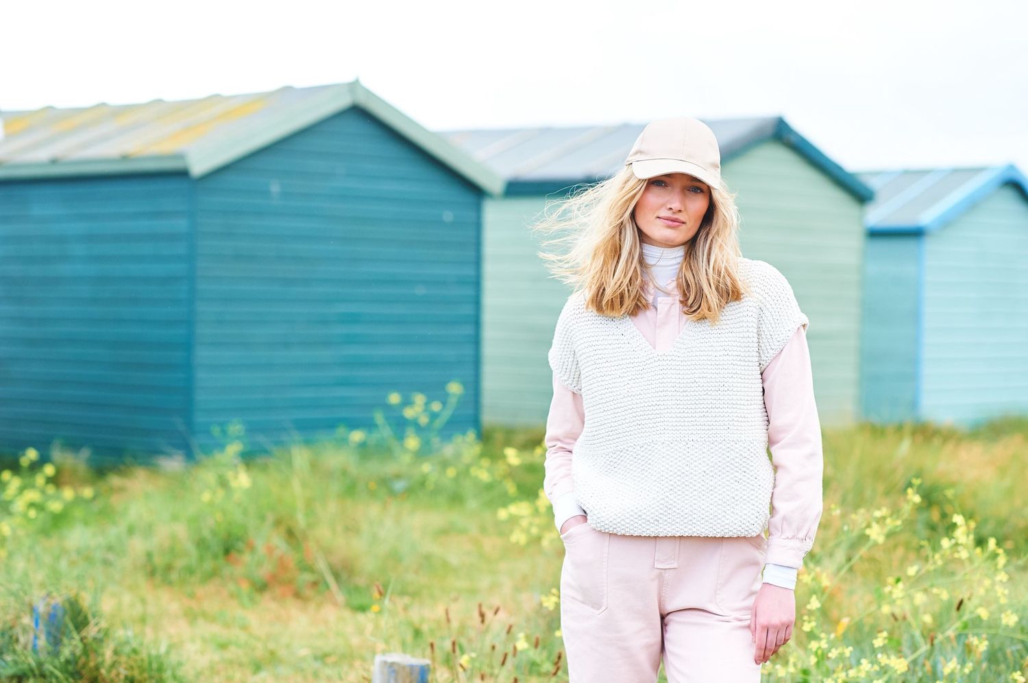 A woman is standing in a field in front of a row of blue beach huts, wearing a cream coloured tank top over a pale pink shirt.