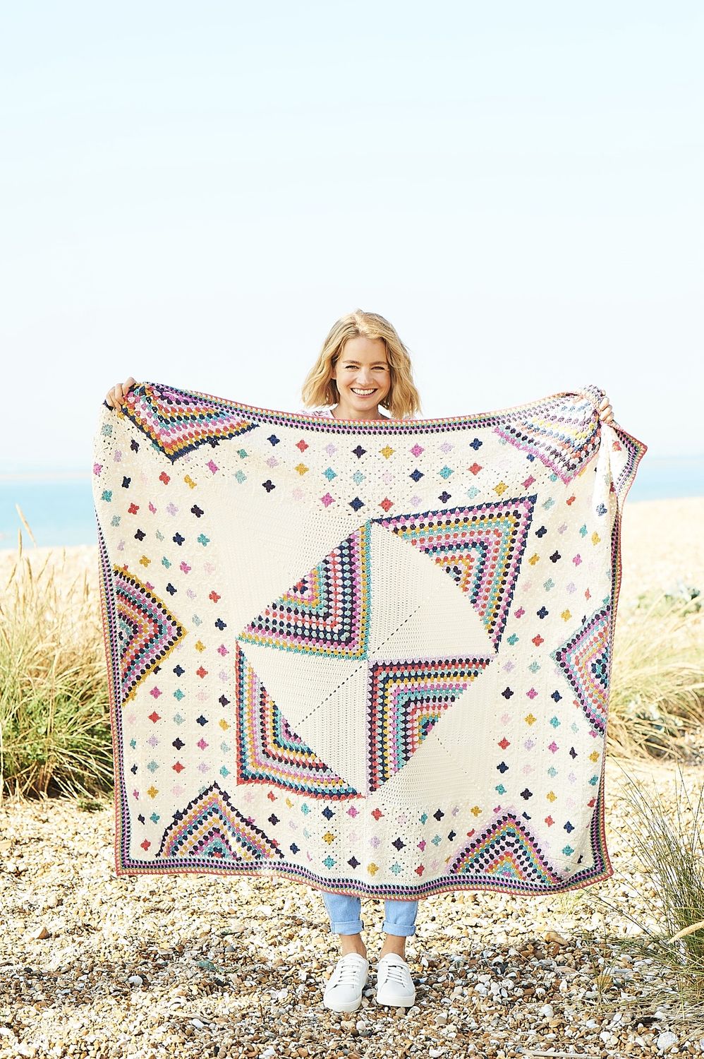 A woman is holding a crocheted blanket made up of squares in cream and various bright colours.