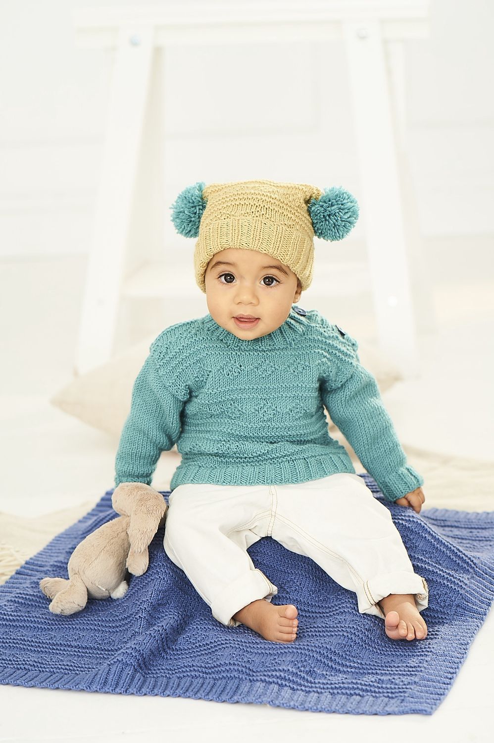 A baby is sitting on a blue knitted blanket, while wearing a light teal knitted sweater and a light green knitted hat with teal pompoms.