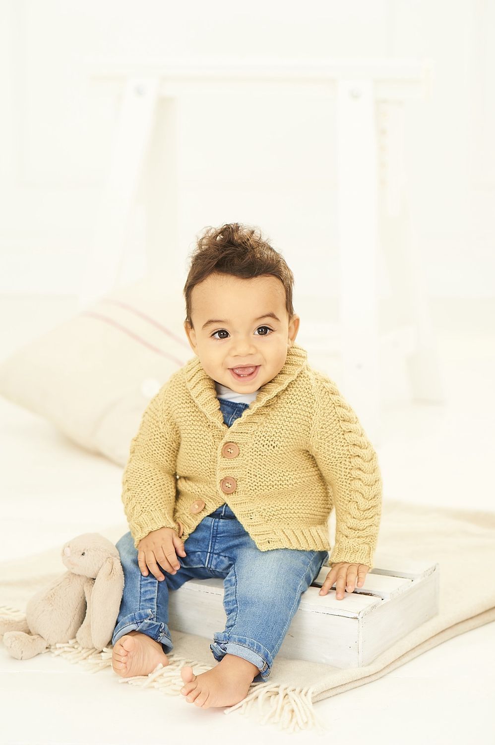 A baby is wearing a yellow knitted cardigan with cable down the sleeves.