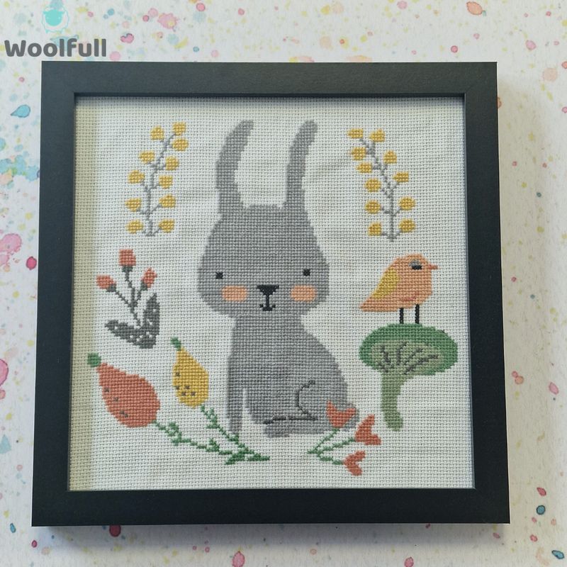 A framed cross stitch with a design of a cartoon style rabbit alongside leaves, flowers and a bird.