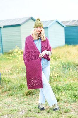 A woman is standing in a field in front of a row of blue beach huts, wearing a green hat and a dark pink shawl.