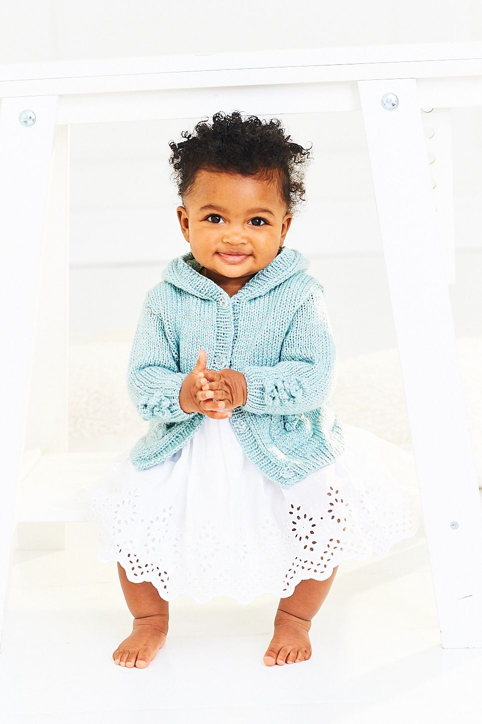 A baby is wearing a blue knitted hooded cardigan with flower detail on the sleeves and pockets.