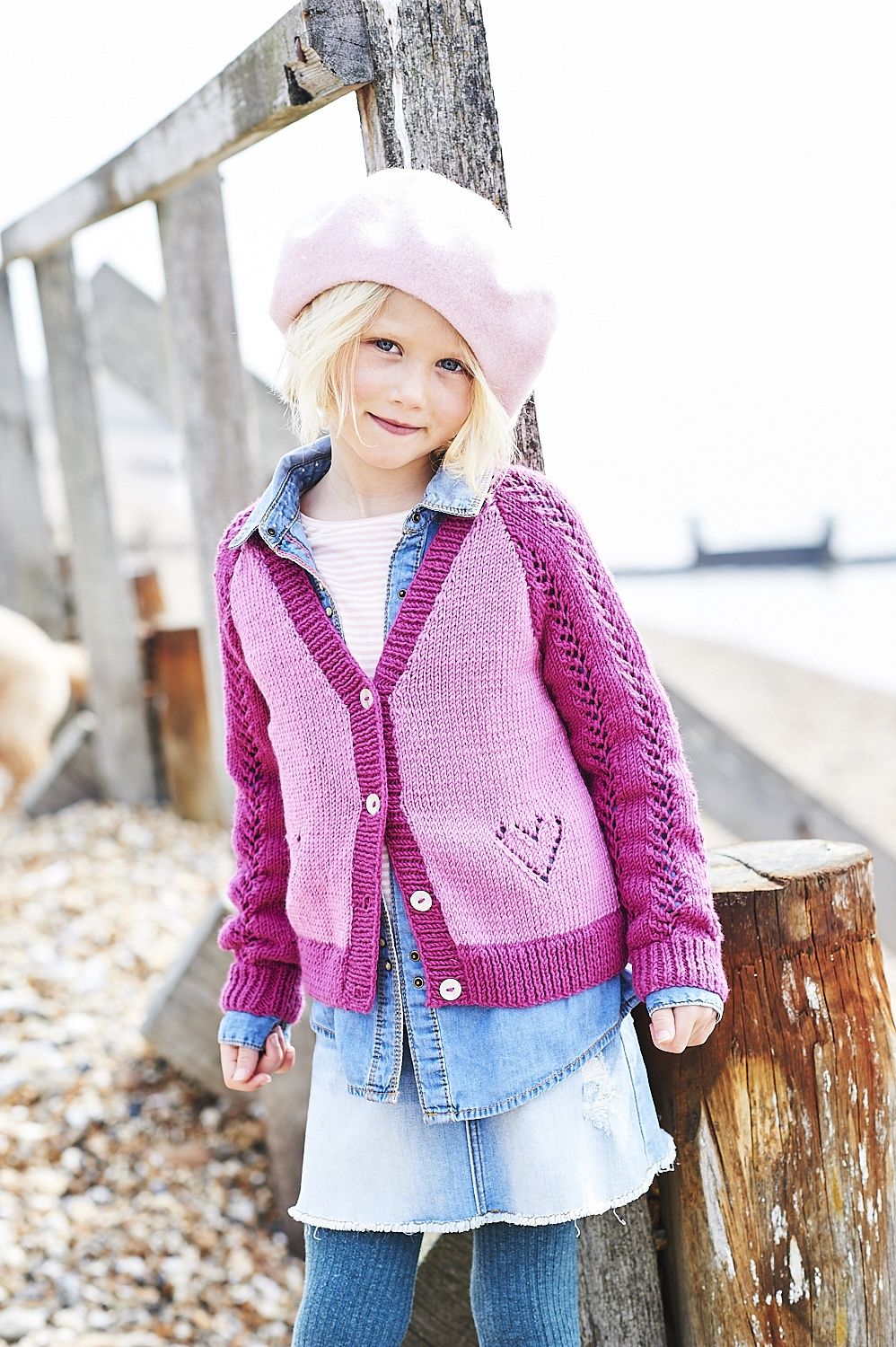 A girl is wearing a light pink knitted cardigan with dark pink sleeves and lace heart detail.