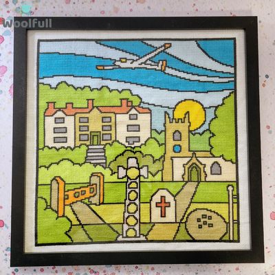 A framed cross stitch with a cartoon style design of the village of Eyam in the Peak District.