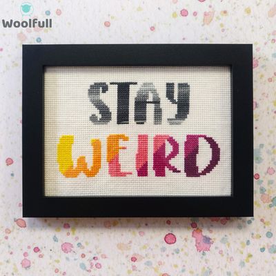 A framed cross stitch with a design that says &#39;stay weird&#39; in grey, yellow, orange, pink and purple.