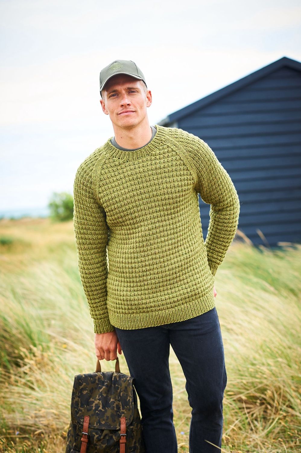 A man is standing in a field in front of a dark blue building, wearing a green textured sweater.
