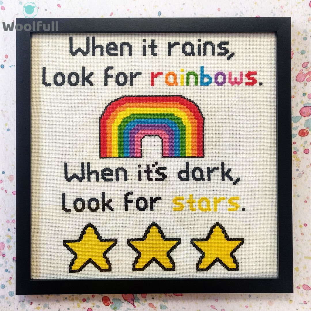 A framed cross stitch with a design that says &#39;When it rains, look for rainbows. When it&#39;s dark, look for stars.&#39; alongside a cartoon style rainbow and 3 yellow stars.