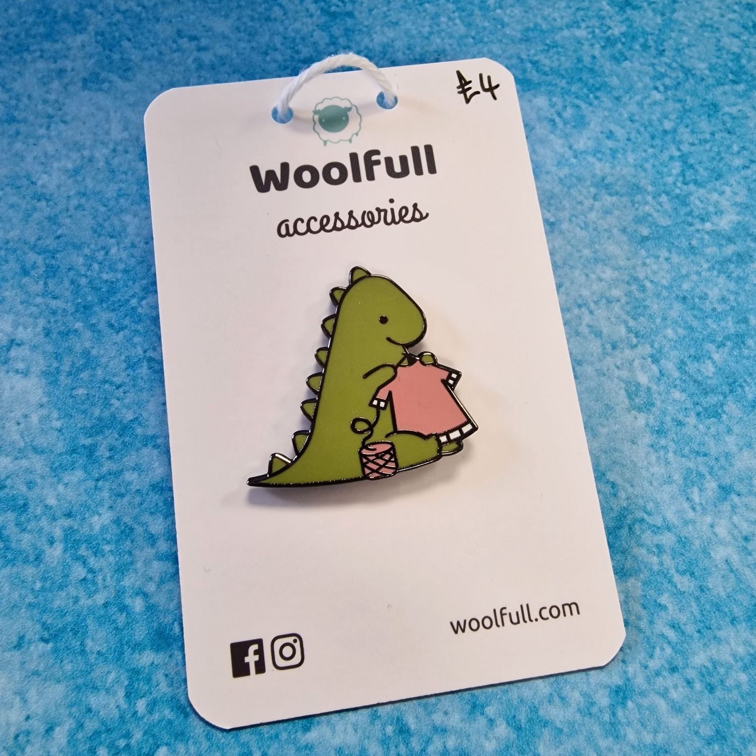 A blue background with a pin badge of a green cartoon dinosaur knitting a pink sweater.