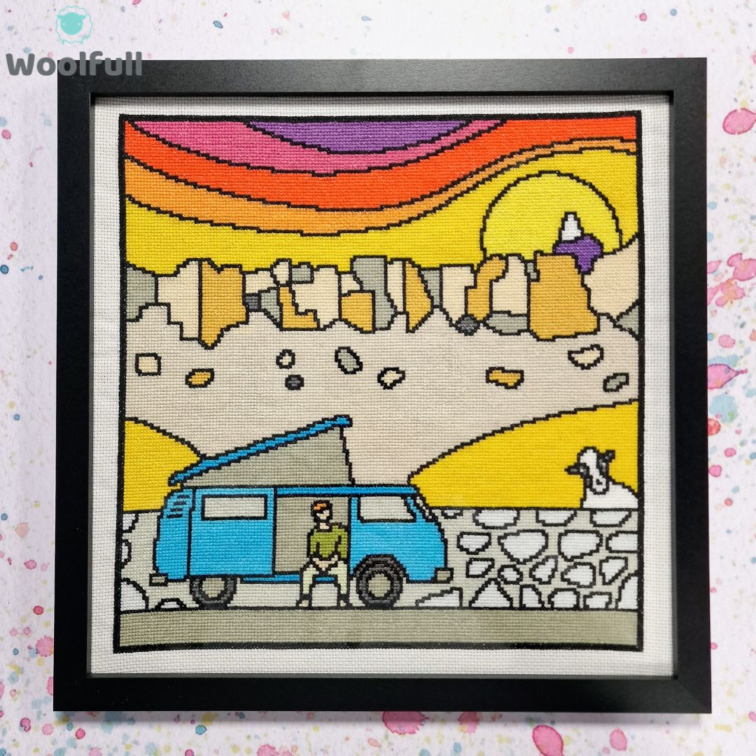 A framed cross stitch with a cartoon style design of Stanage Edge in the Peak District with a blue camper van in front.