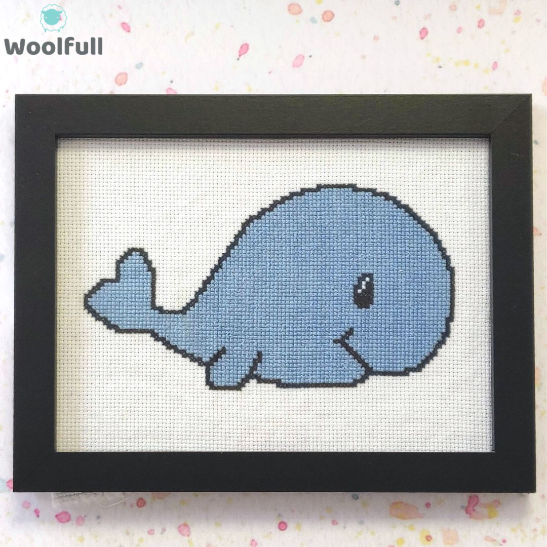 A framed cross stitch with a design of a blue cartoon style whale.