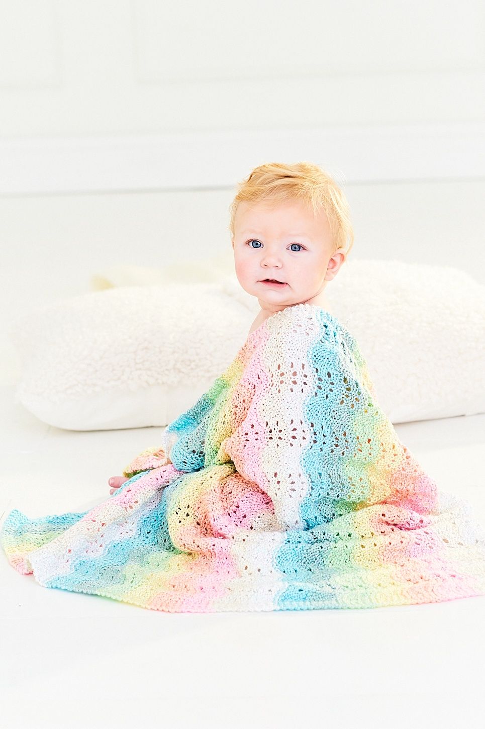 A baby has a knitted ripple blanket in white, cream, pink, blue, green, yellow and orange draped around their shoulders.