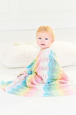 A baby has a knitted ripple blanket in white, cream, pink, blue, green, yellow and orange draped around their shoulders.
