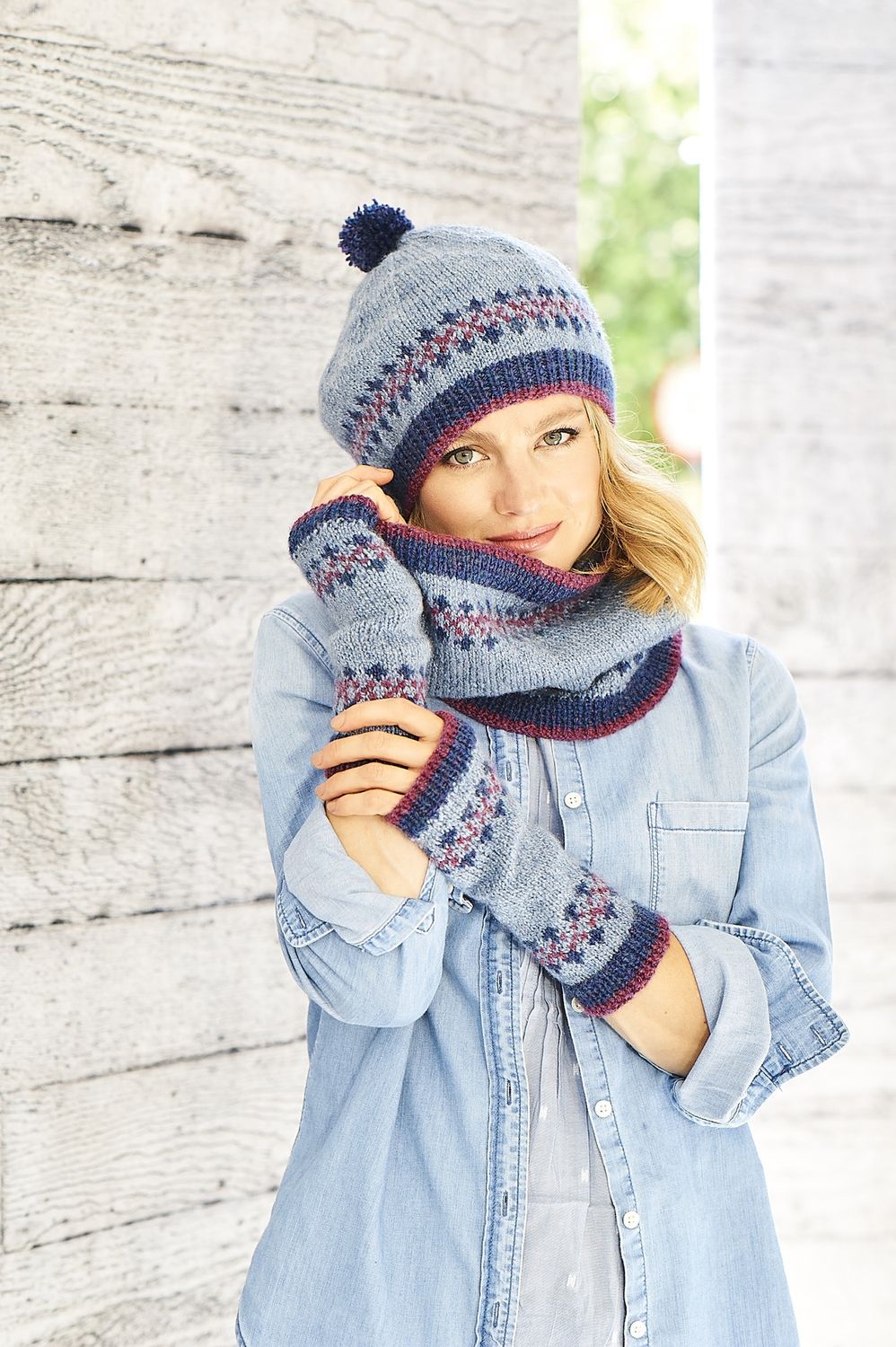 A woman is wearing a knitted hat, cowl and wristwarmers, all in light blue with small amounts of pink and dark blue colourwork.