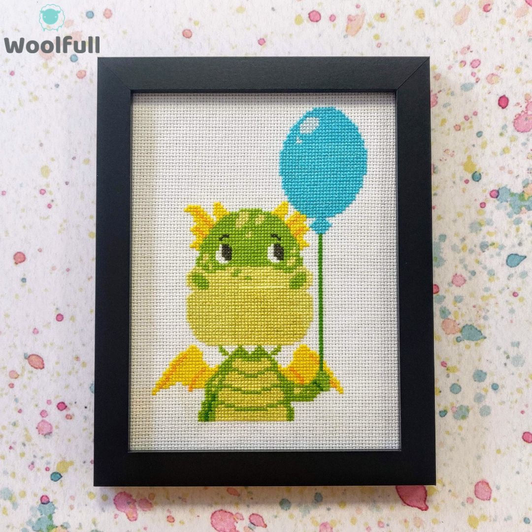 A framed cross stitch with a design of a green cartoon dragon holding a blue balloon.