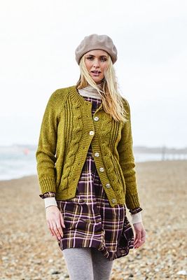 A woman on the beach is wearing a short green cardigan with cable details down the front.