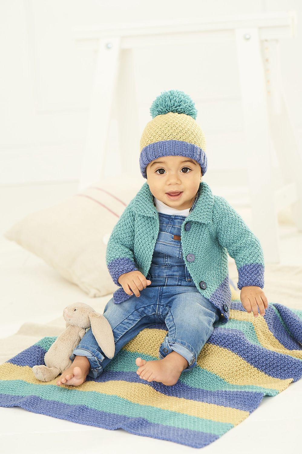 A baby is sitting on a blue, teal and green stripy knitted blanket while wearing a matching knitted cardigan and hat.