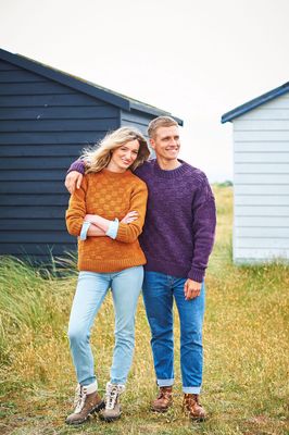 A woman and a man are standing in a field in front of a dark blue building. The woman is wearing an orange sweater and the man is wearing a dark purple sweater. Both sweaters have a textured checkerboard pattern.