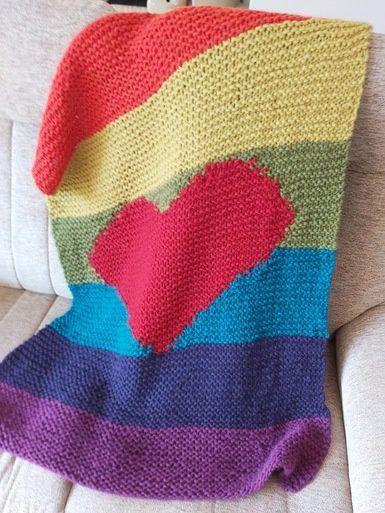 A knitted blanket in orange, yellow, green, blue and purple stripes, with a red heart in the centre.