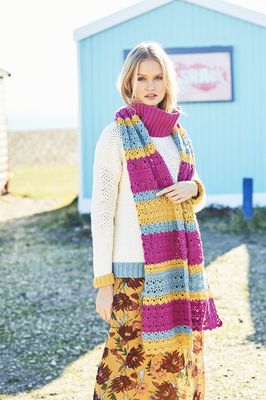 A woman is wearing a pink, blue and yellow crocheted scarf on top of a cream crocheted sweater with pink, blue and yellow cuffs.