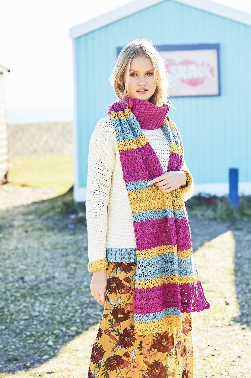 A woman is wearing a pink, blue and yellow crocheted scarf on top of a cream crocheted sweater with pink, blue and yellow cuffs.