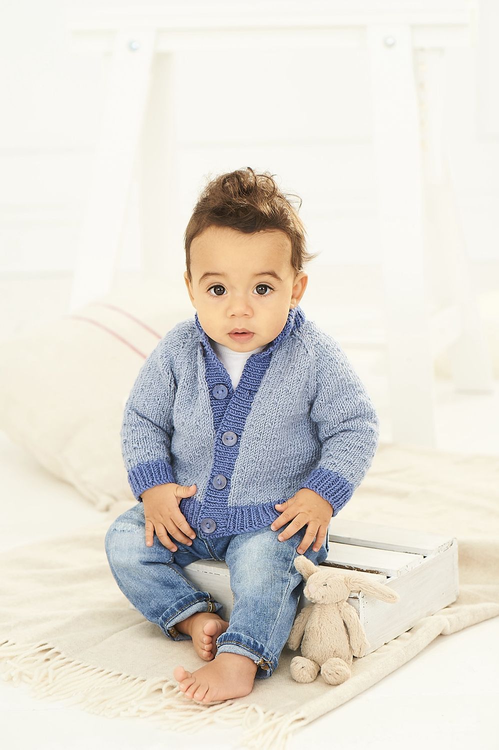 A baby is wearing a light blue knitted cardigan with darker blue cuffs and button band.